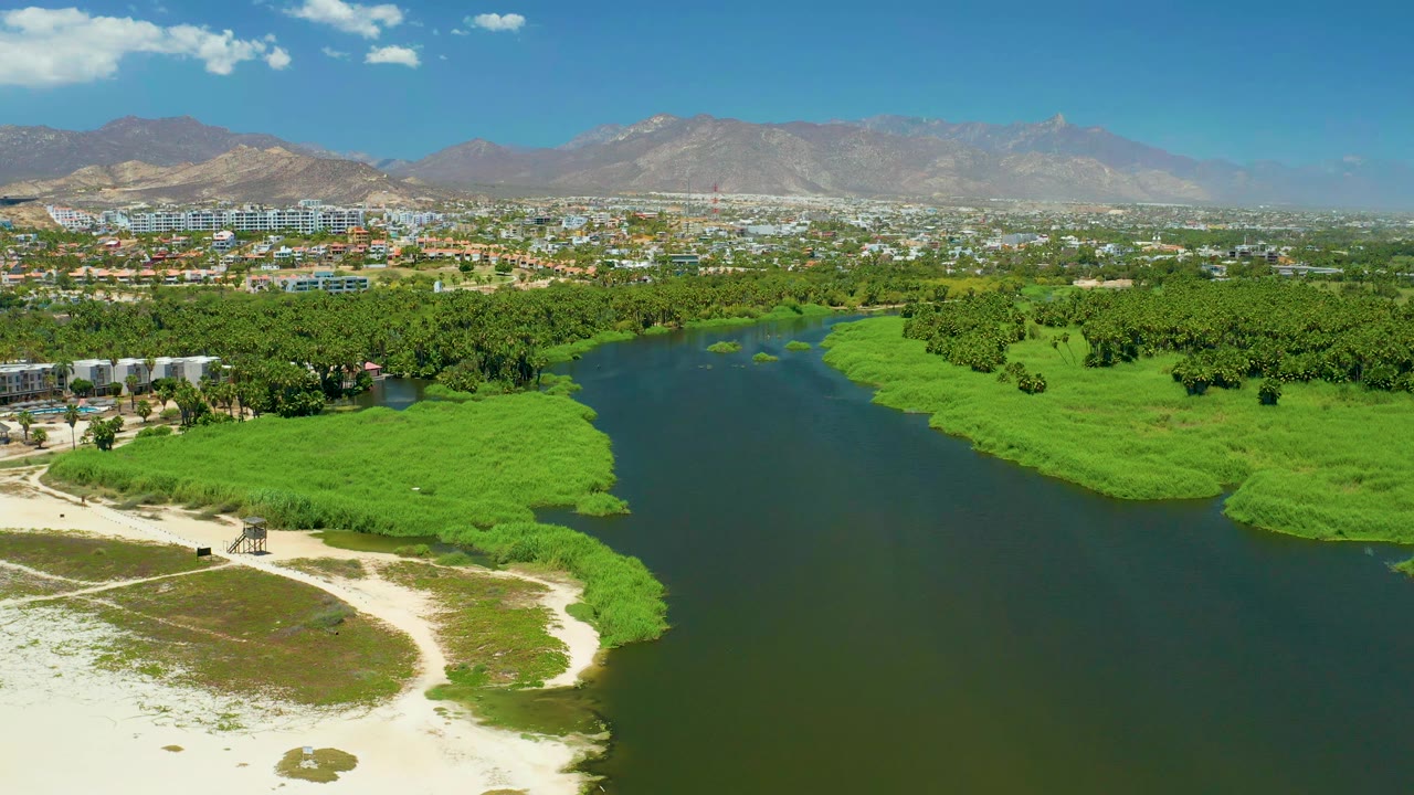 Wide aerial frame of San Jose del Cabo Estuary showing the river channel slightly farther across the green banks, with sandy shore at lower left, distant city buildings, mountains, and no visible people.