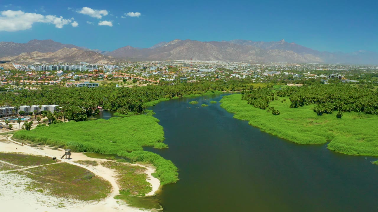 Mid-clip aerial view of San Jose del Cabo Estuary with the river bordered by dense green vegetation, pale sand in the lower foreground, city buildings along the far edge, hazy mountains, and no visible people.