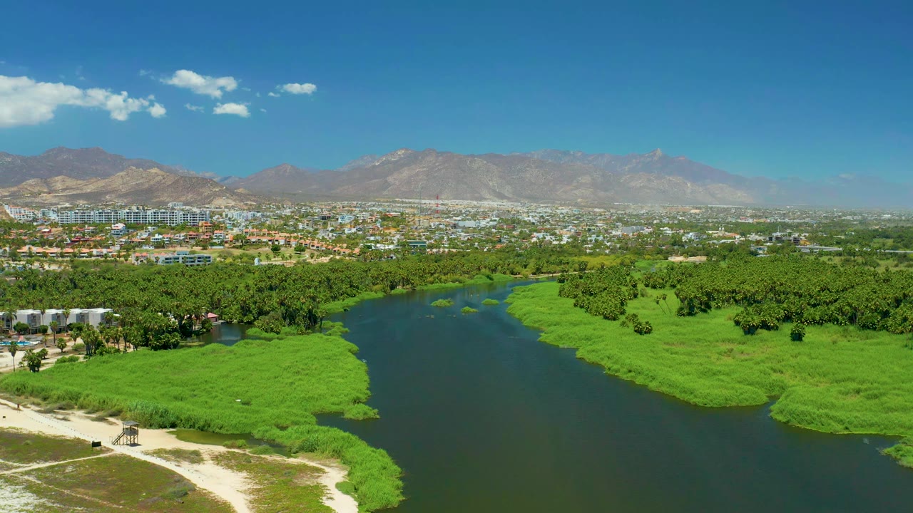 Aerial frame over San Jose Del Cabo Estuary showing the river channel slightly farther across the lower center, with dense green banks, low city blocks, mountain ridges, blue sky, clouds, and no visible people.