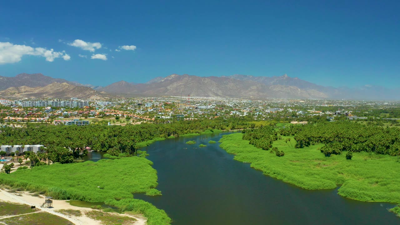 Mid-clip aerial over San Jose Del Cabo Estuary with the river bend placed left of center, green marsh and tree canopy filling the foreground, city buildings beyond, mountains on the horizon, and no visible people.