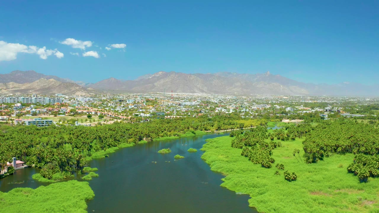 Opening aerial frame of the San Jose Del Cabo Estuary, with a dark river bordered by bright marsh grass, dense trees, distant city buildings, hazy mountains, blue sky, and no visible people.
