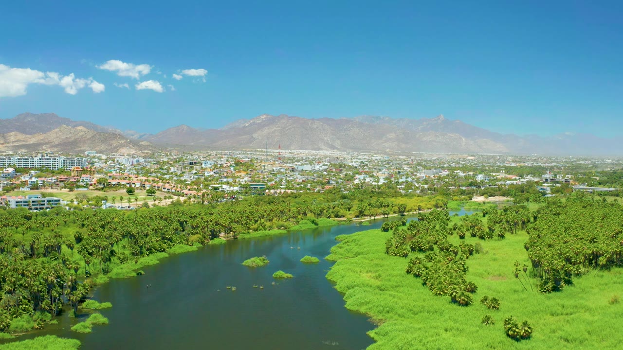 Second frame holds the San Jose del Cabo Estuary from above, with the river curving past dense green vegetation toward low buildings and mountains, with no visible people.