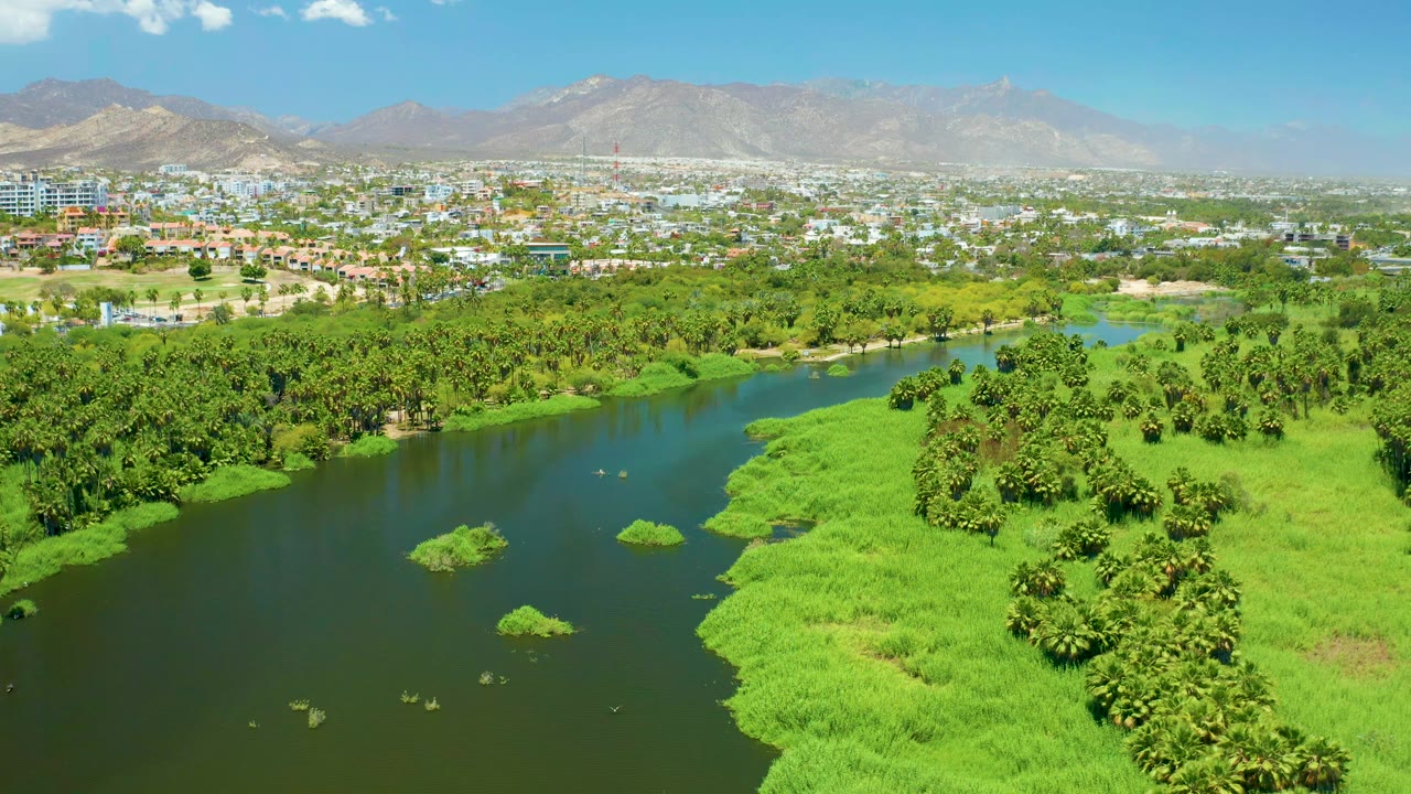 Wide aerial over a river bend with bright green banks and distant buildings.