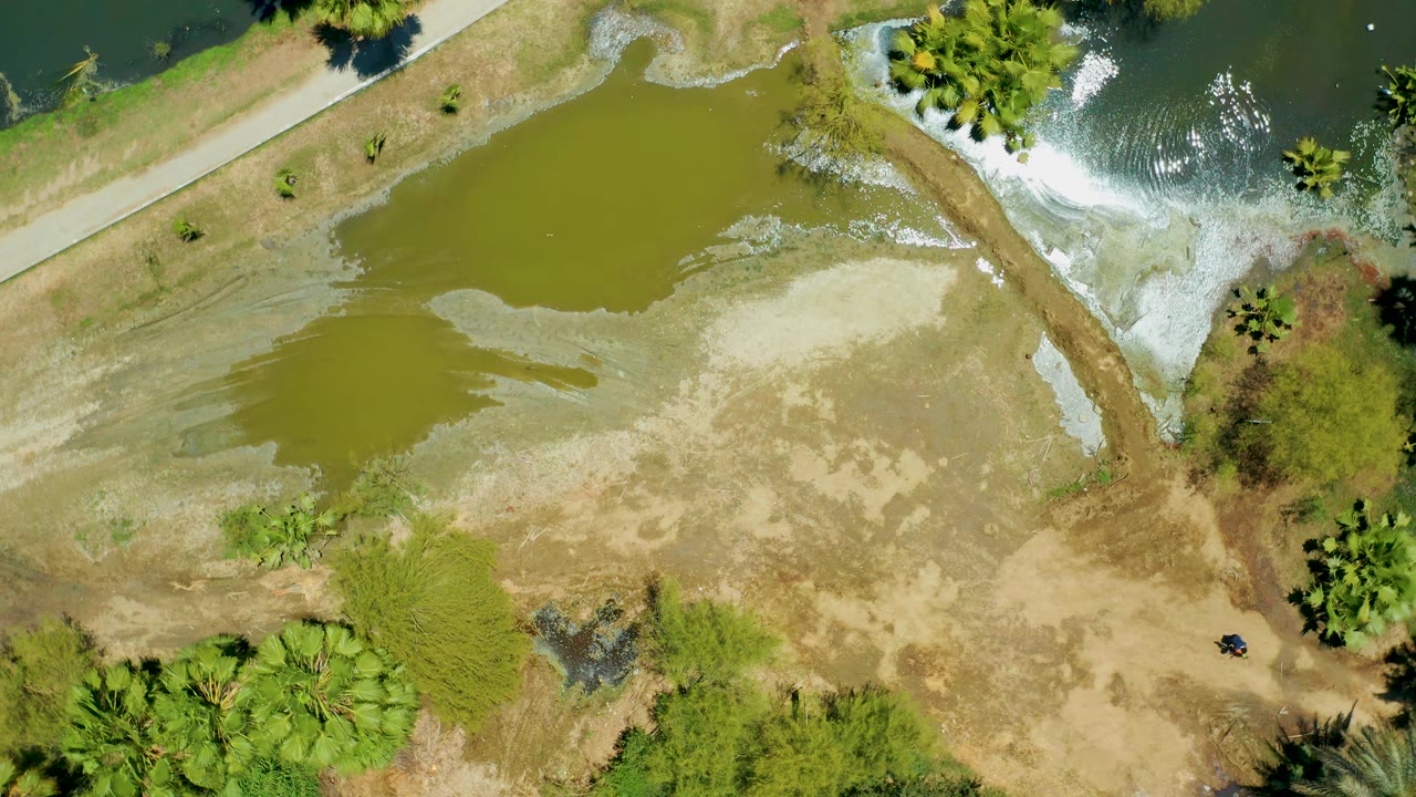 Second overhead frame at San Jose del Cabo Estuary shows the pond shifted slightly in view, pale exposed mudflats, river water with white ripples, a road or path, trees, and no visible people.