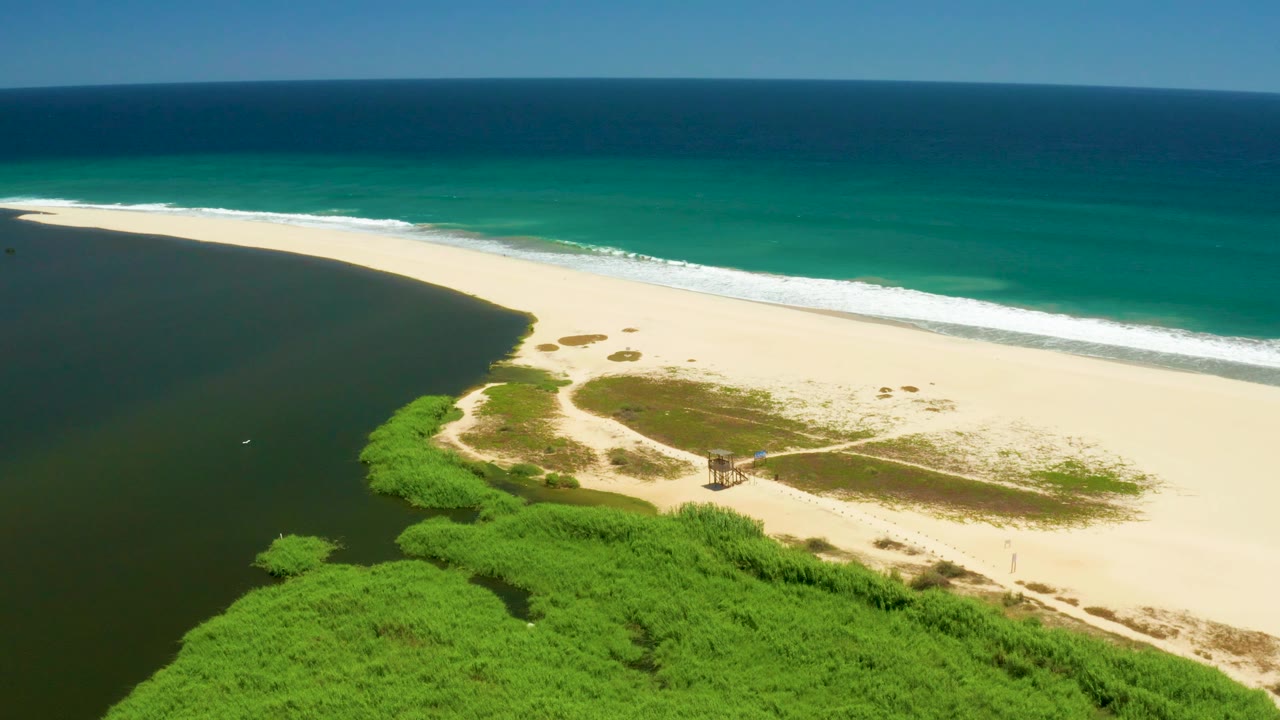 San Jose Del Cabo Estuary aerial frame with more open dark inlet water on the left, a long pale beach across the center, low dunes or vegetation patches on the sand, ocean surf beyond, and no visible people.