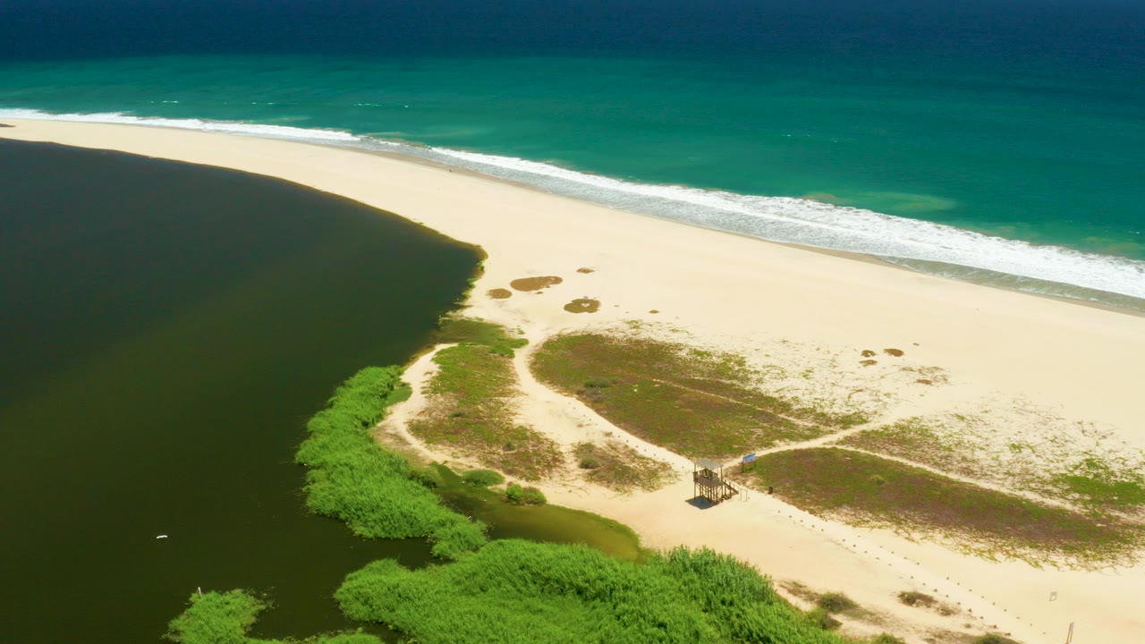 High aerial view over San Jose Del Cabo Estuary showing dark lagoon water beside pale sand, green vegetation, turquoise surf, a small beach hut near a path, and no visible people.
