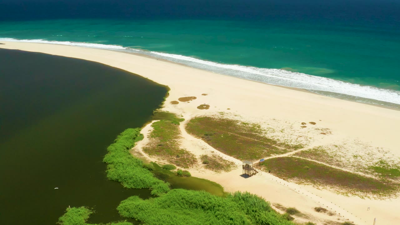 Aerial frame of the San Jose Del Cabo Estuary shoreline with the lagoon filling the left side, white waves along the upper beach, green vegetation below, the hut near a sandy path, and no visible people.