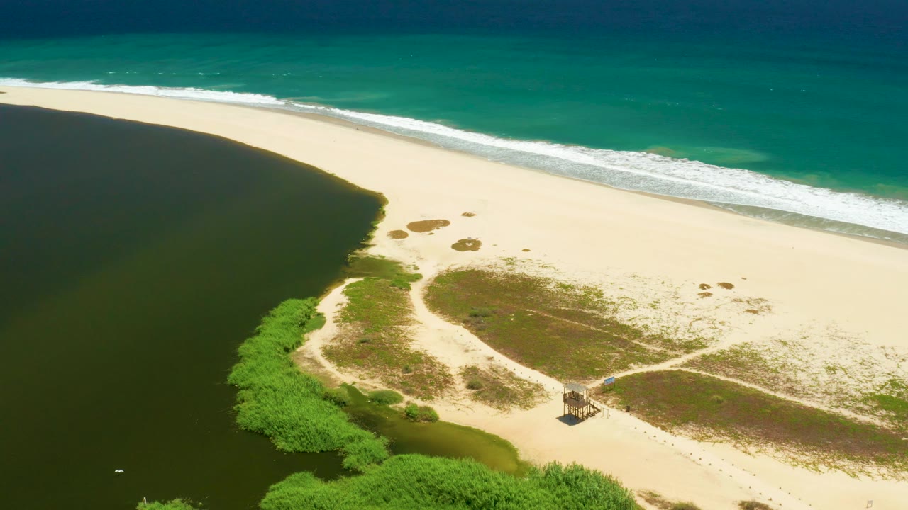 Aerial view in San Jose Del Cabo Estuary with dark lagoon water curving along the sand, bright beach and grass patches in the middle, ocean surf at the top, a small hut near the lower path, and no visible people.