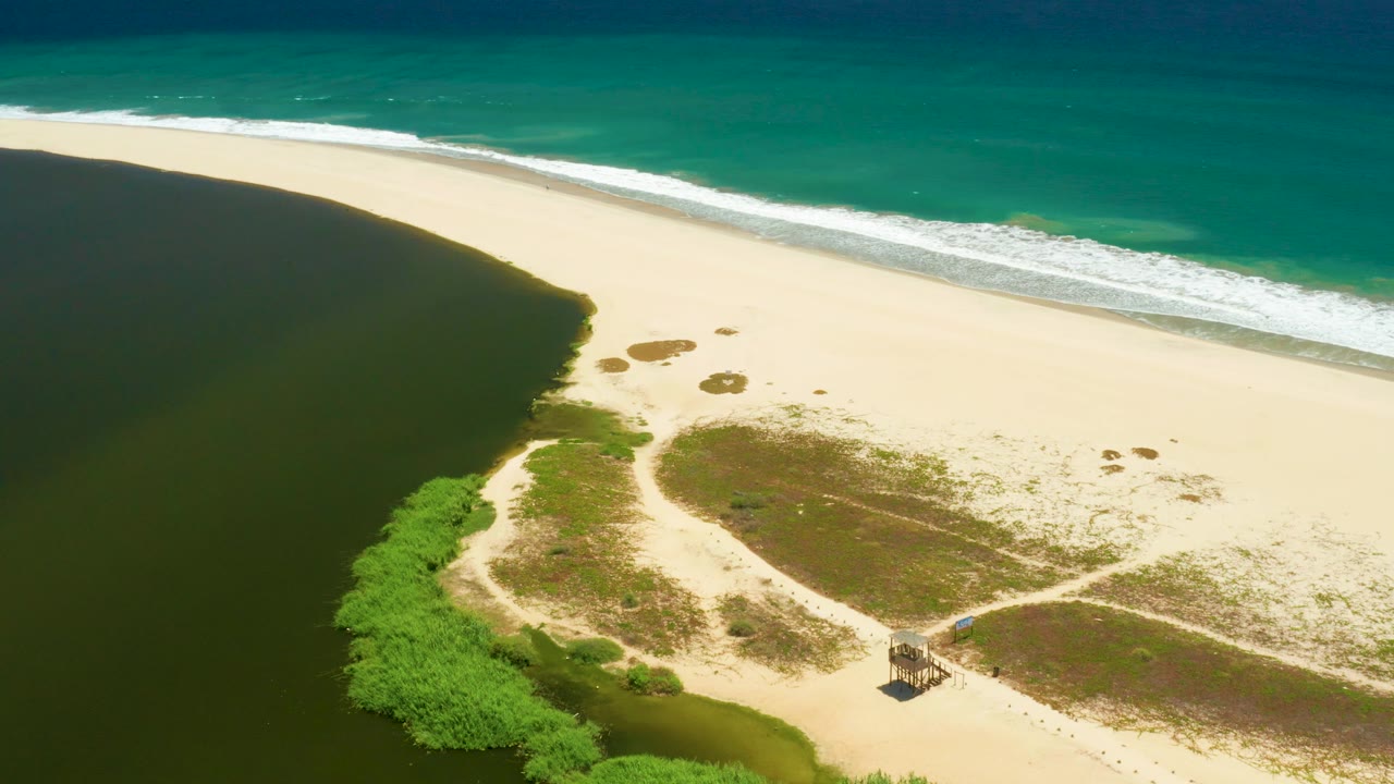 Aerial beach and lagoon view with hut, vegetation, sand patches, and ocean waves.
