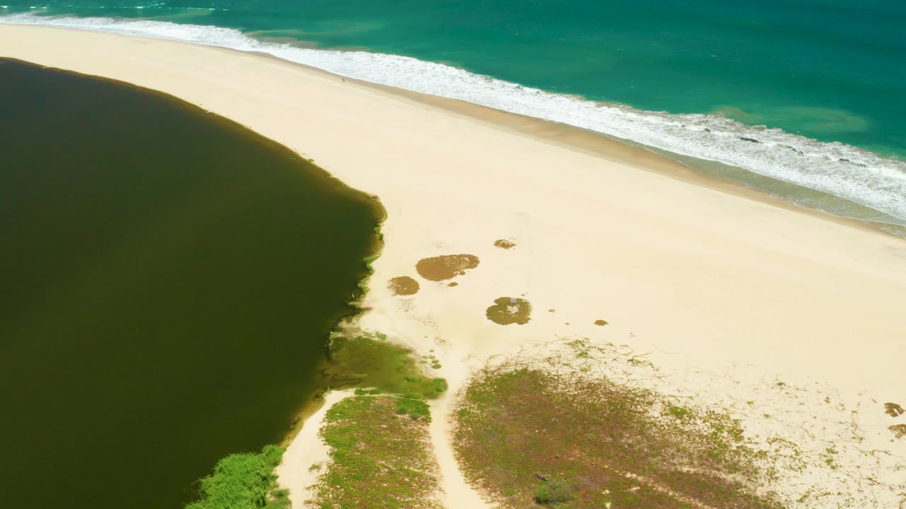 At 0:03, the aerial view of San Jose Del Cabo Estuary shows more of the lagoon edge at left, scattered wet patches on the sand, and ocean foam tracking along the top, with no visible people.