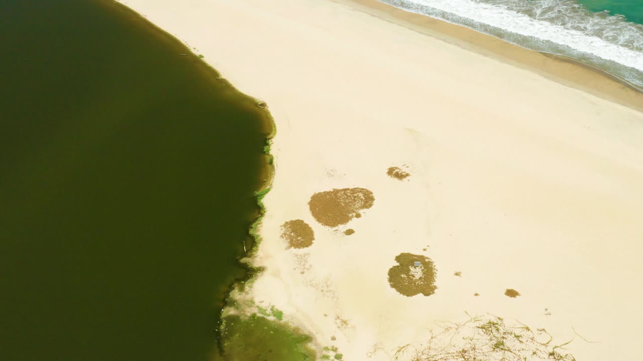 San Jose Del Cabo Estuary shoreline seen from above, with the tidal pool’s curved dark edge on the left, bright sand across the frame, waves along the upper right, and no visible people.