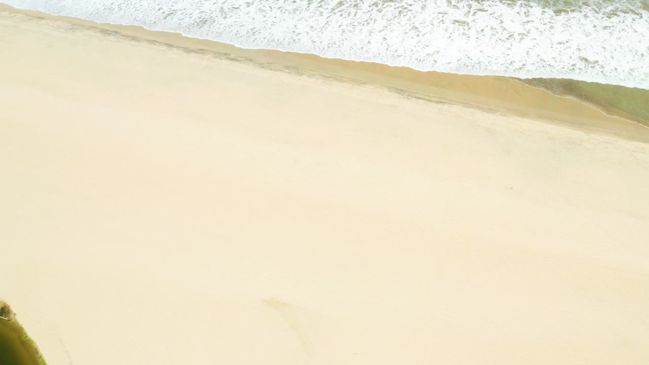 Aerial view of San Jose Del Cabo Estuary beach with a broad pale sand field, foamy surf at the top of frame, a small dark green edge near the lower left, and no visible people.