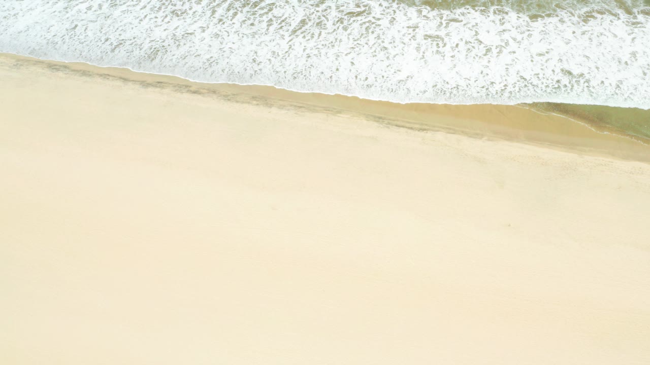 Aerial frame over San Jose Del Cabo Estuary beach where white ocean foam spreads across the upper half and pale sand fills the lower half, with no visible people.