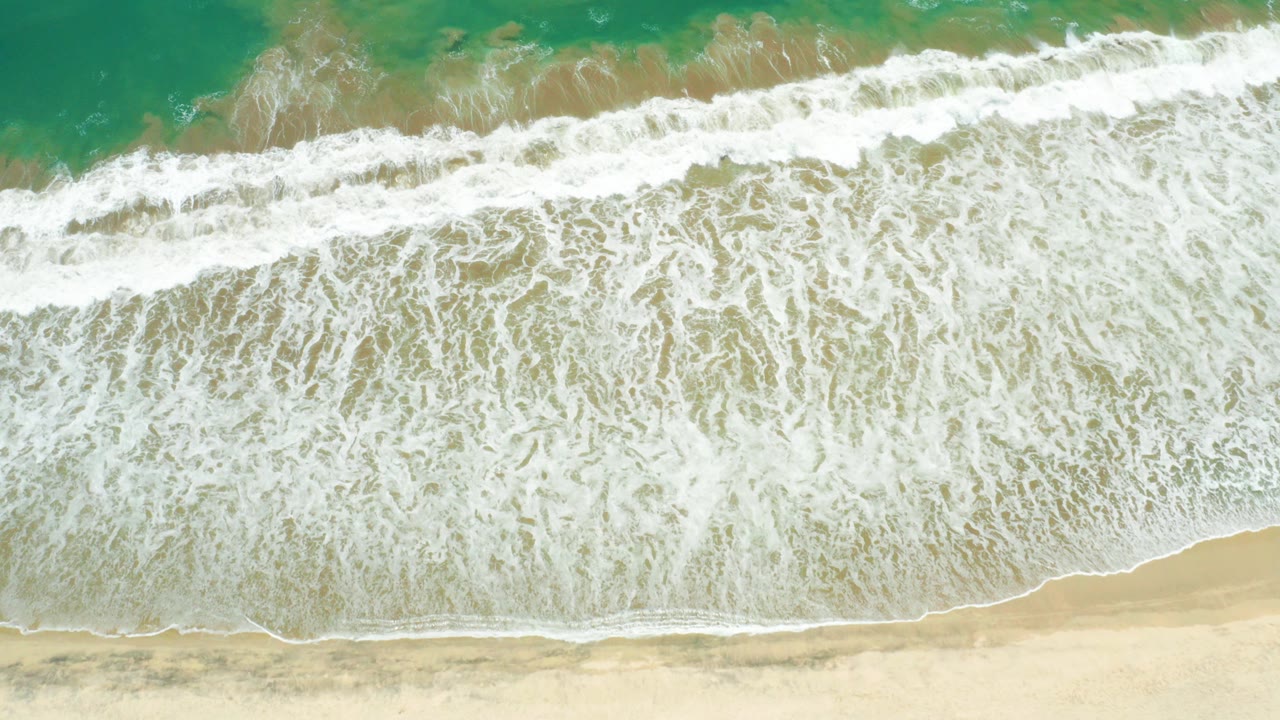 Overhead view at San Jose Del Cabo Estuary showing white surf spread across pale sand with turquoise water above and no visible people.
