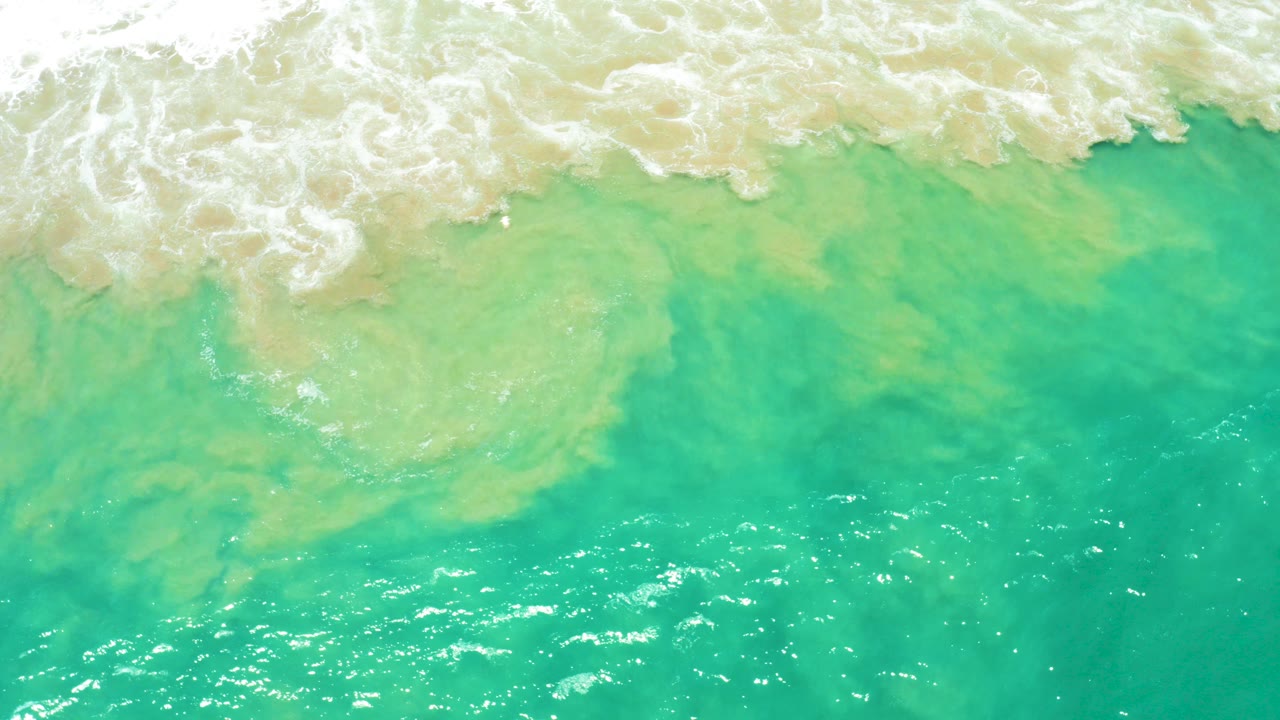 Overhead view of turquoise ocean water near San Jose Del Cabo Estuary, with pale sandy shallows and white foam entering along the top edge, no visible people.