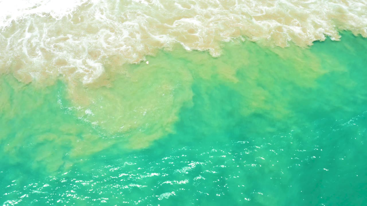Aerial frame over San Jose Del Cabo Estuary ocean water, where white foam spreads across the upper left and green turquoise water fills the lower frame, no visible people.