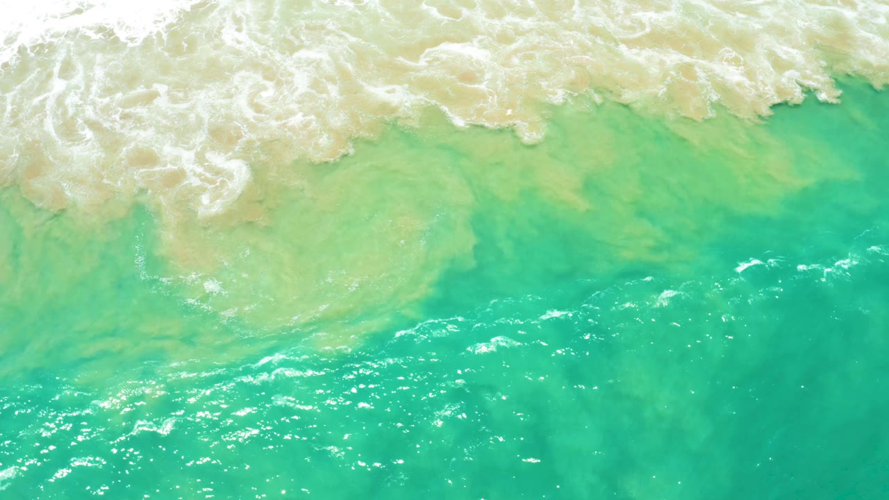 Overhead San Jose Del Cabo Estuary ocean view with a broad white foam band across the top and brighter turquoise water with small ripples below, no visible people.