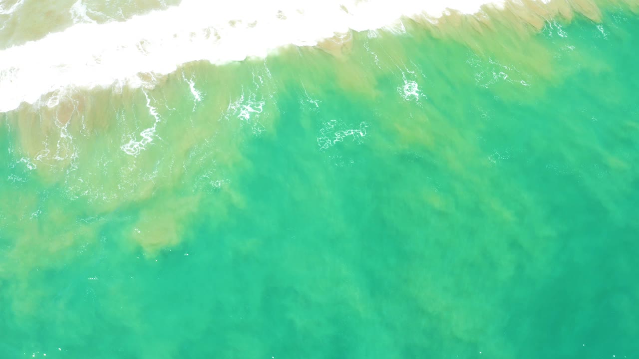 Overhead frame from San Jose Del Cabo Estuary with white surf pushed toward the upper edge and a wider field of clear turquoise water below, with no visible people.