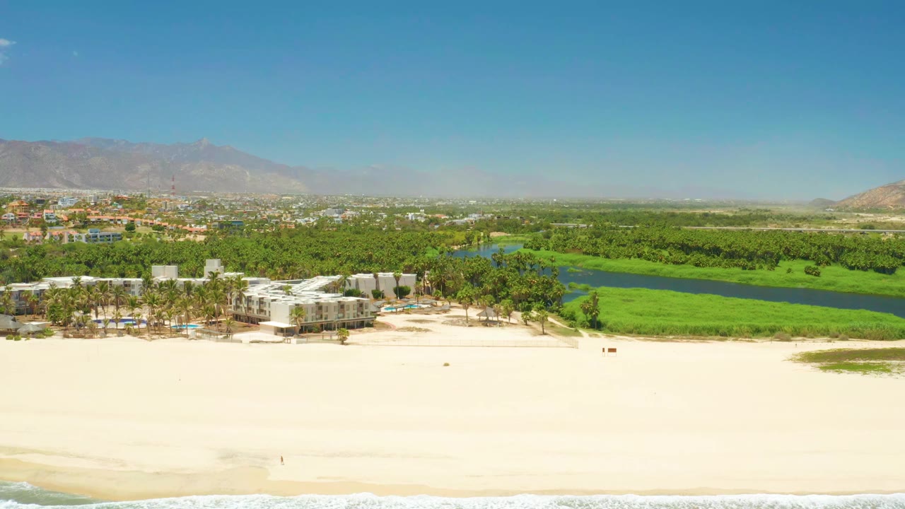 Opening aerial frame over San Jose Del Cabo Estuary with pale sand in front, a blue-green lagoon behind palm trees and buildings, distant mountains under clear sky, and no visible people.