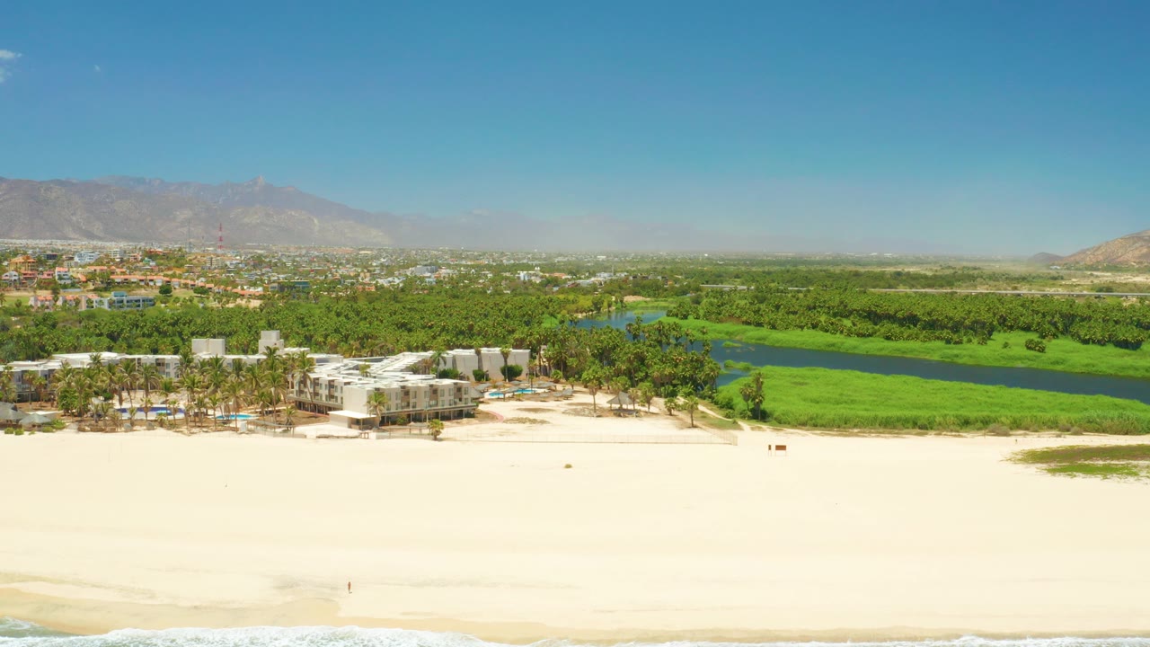 Aerial view across empty-looking beach toward palms, lagoon water, buildings, and mountains.