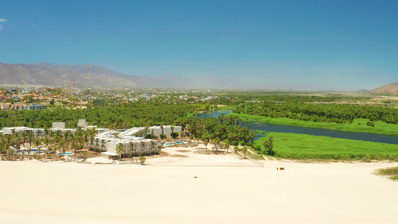 Opening aerial frame over San Jose del Cabo Estuary, with pale sand below, white beachside buildings and palms near the lagoon, distant mountains, blue sky, and no visible people.