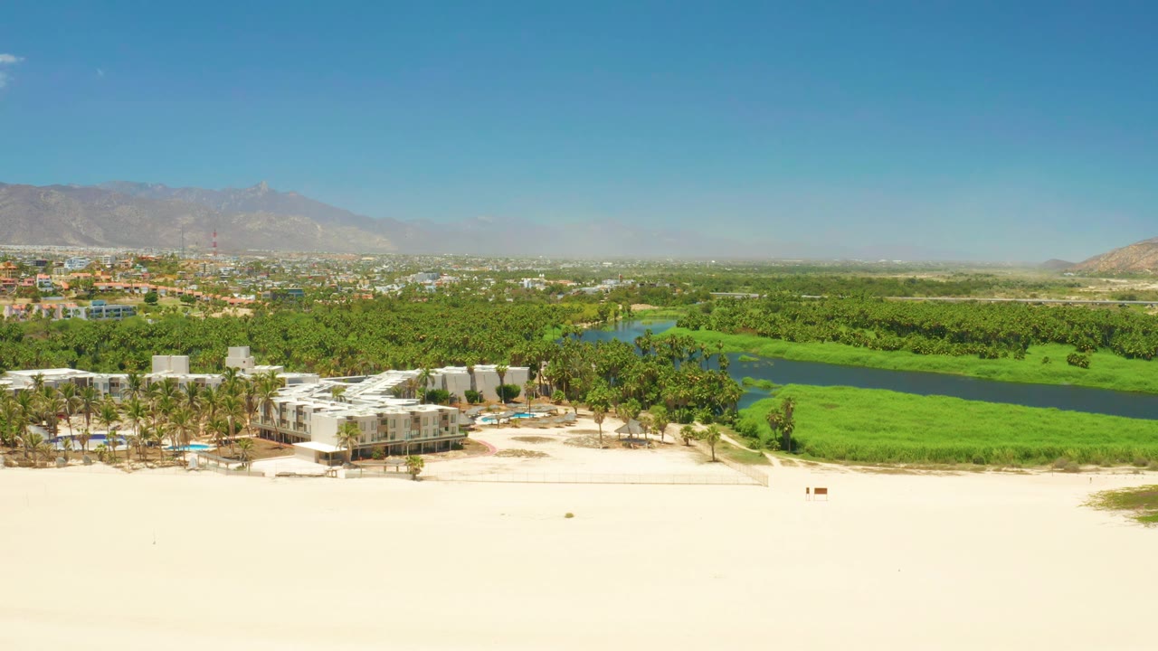 San Jose del Cabo Estuary aerial view with the white coastal buildings slightly left of center, the green lagoon stretching behind them, hazy mountains on the horizon, and no visible people.