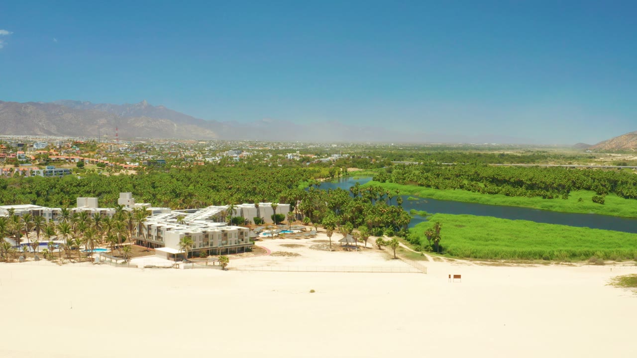 San Jose del Cabo Estuary seen from above, with broad sand in the foreground, palm-lined white buildings near the lower center, lagoon water and greenery behind, and no visible people.