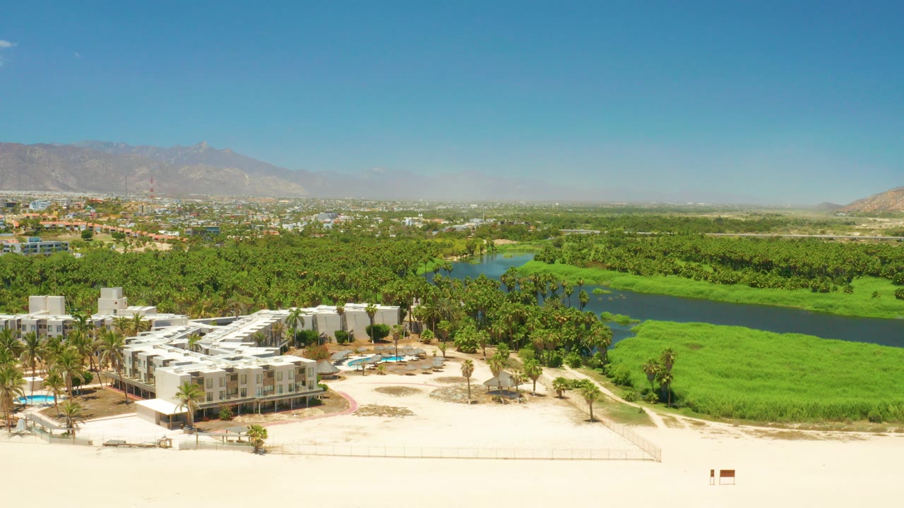 Opening aerial frame over San Jose del Cabo Estuary with white resort buildings and sandy beach in the foreground, green lagoon banks and dark water beyond, distant mountains, and no visible people.