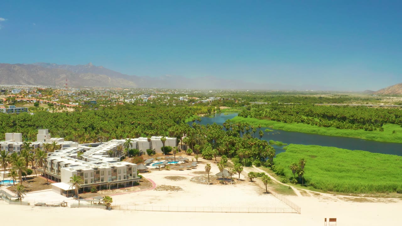 Wide San Jose del Cabo Estuary view with white resort blocks at lower left, a bright green lagoon corridor across the middle, dark water curves, distant settlement, mountains, and no visible people.