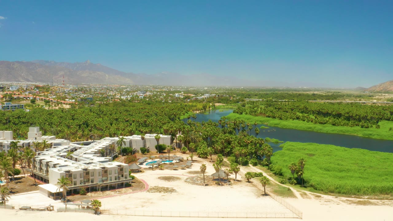Aerial resort foreground with lagoon, palms, city edge, and mountain backdrop
