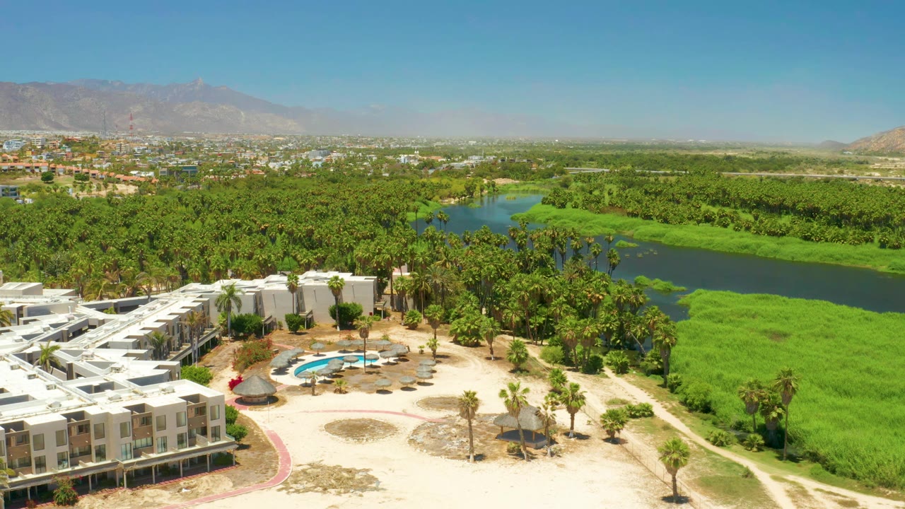 Elevated frame over San Jose Del Cabo Estuary with the white resort block still prominent at lower left, a curved pool below, green water through the center, palms, mountains, and no visible people.