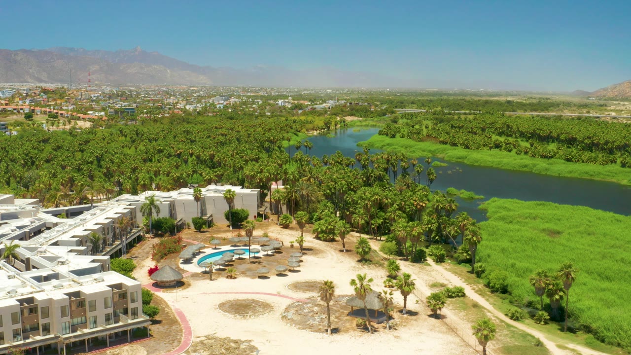 Aerial frame from the San Jose Del Cabo Estuary clip with resort roofs at lower left, a blue pool near center bottom, palm grove, green river bends, distant developed area, mountains, and no visible people.