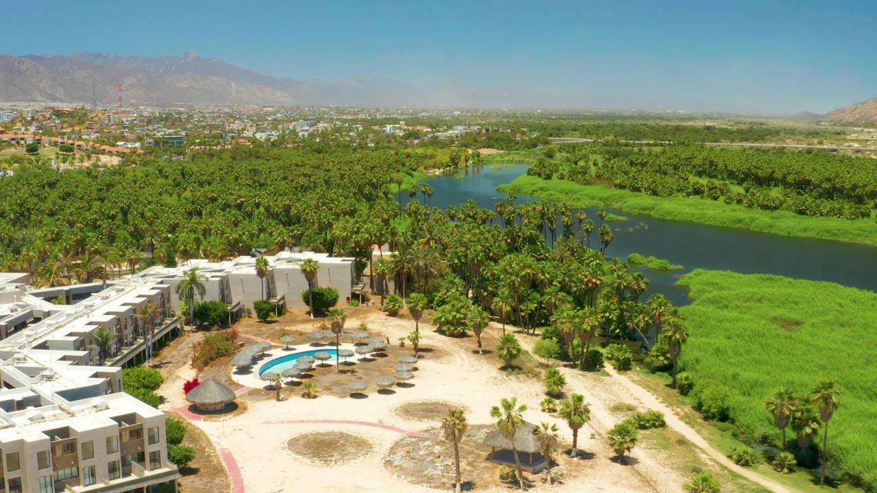 Mid-clip aerial over San Jose Del Cabo Estuary showing the resort sliding farther toward the lower left, the blue pool in sandy grounds, thick palms, green river water, mountains, and no visible people.