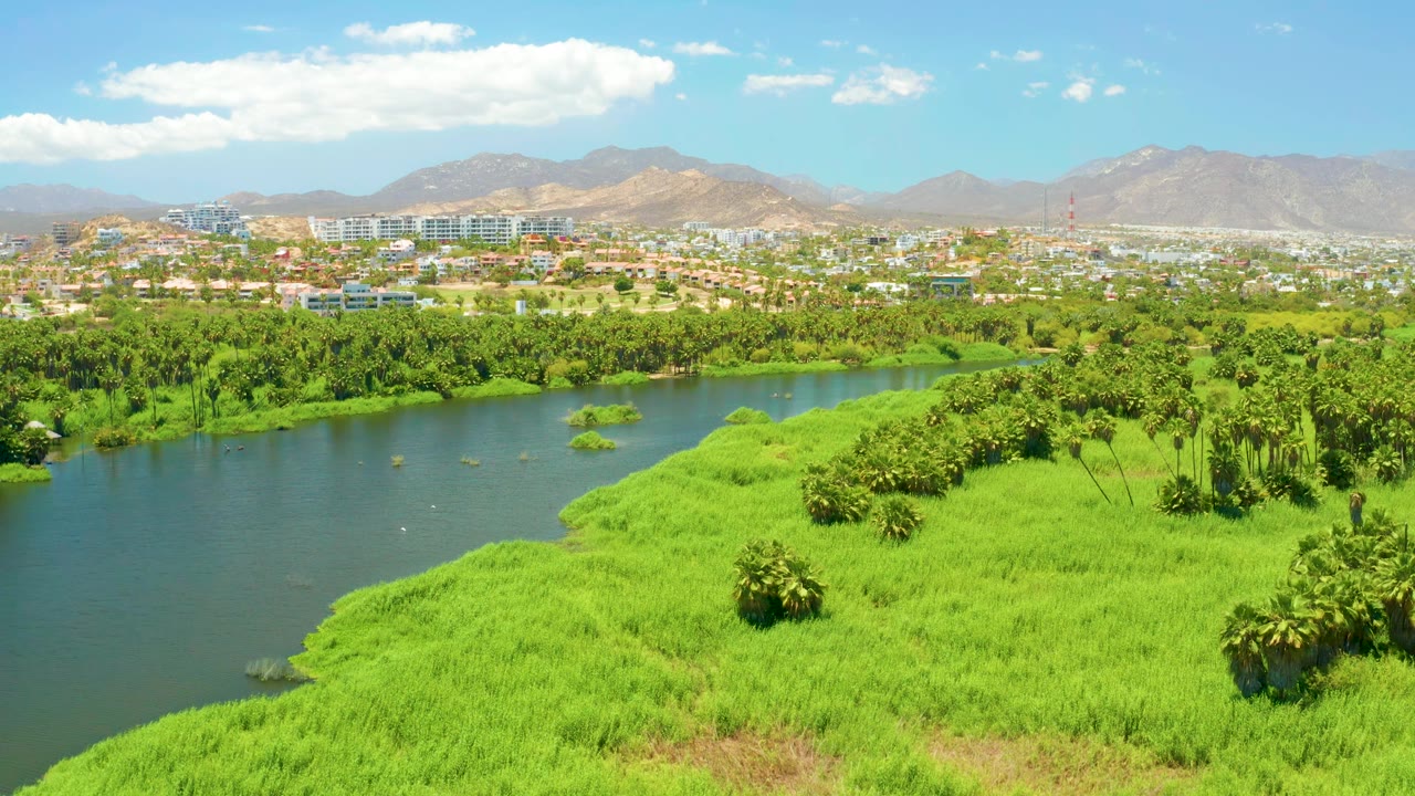 Aerial frame over San Jose Del Cabo Estuary with the river running along the left side, marshy green banks across the lower frame, distant buildings, mountains, scattered clouds, and no visible people.