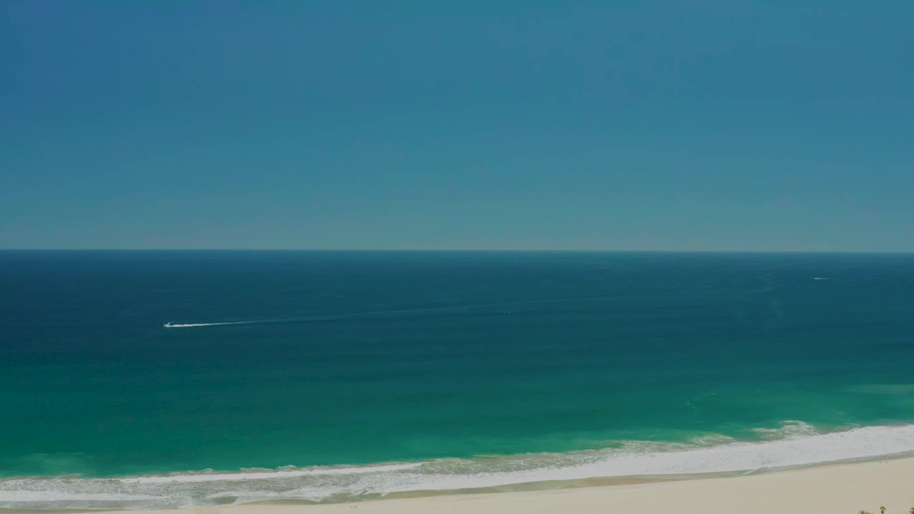 Opening frame of San Jose Del Cabo Estuary coastline with blue sky, turquoise water, white surf on pale sand, a small distant boat wake, and no visible people.