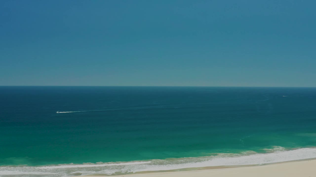 San Jose Del Cabo Estuary shoreline frame with the boat wake farther across the blue water, white breakers approaching the sand, clear sky above, and no visible people.