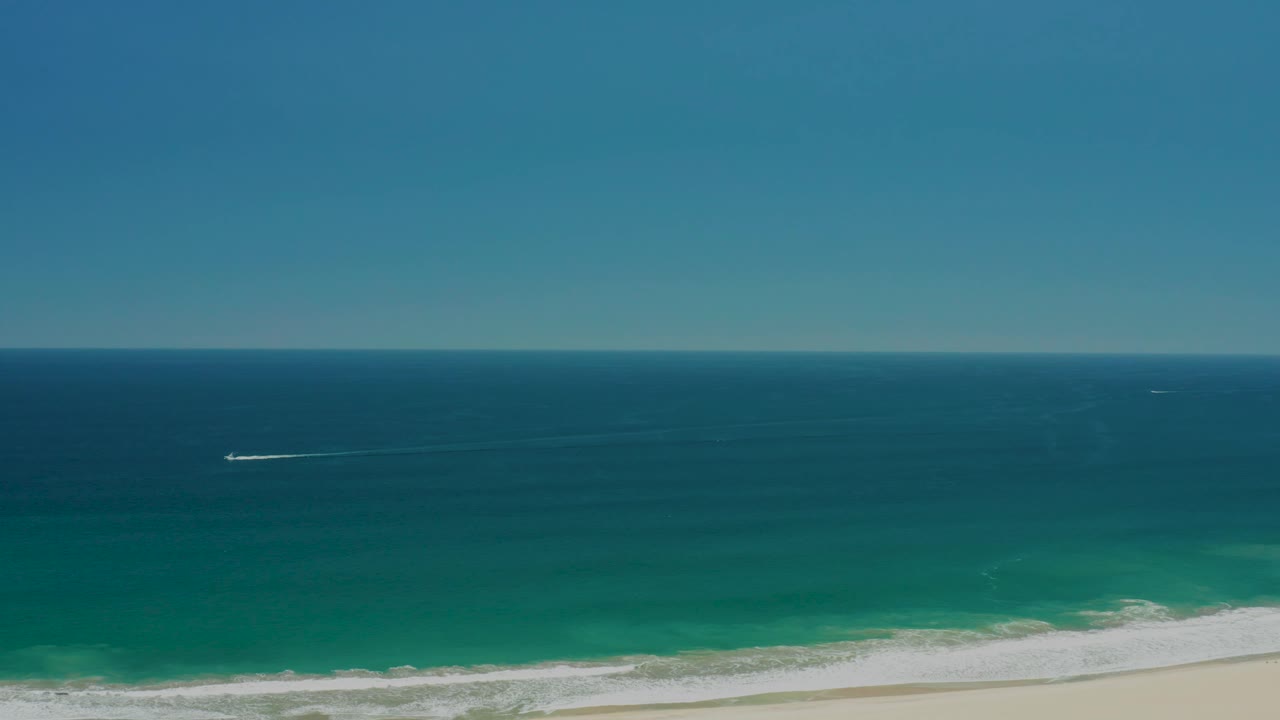 Mid-clip aerial frame over San Jose Del Cabo Estuary coast, with a large band of sky, dark blue ocean, green shallows, white surf, sandy beach, a small boat wake, and no visible people.