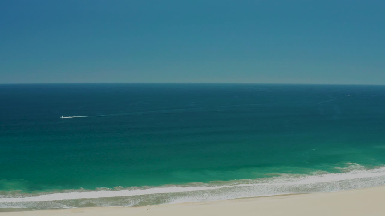 Aerial frame over San Jose Del Cabo Estuary coastline showing deep blue ocean, turquoise water near shore, white surf, pale sand, distant boat wakes, and no visible people.