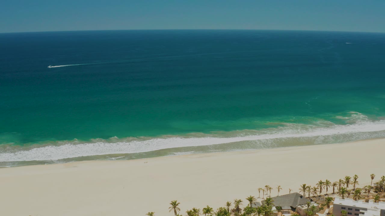 Aerial frame over San Jose del Cabo Estuary beach with deep blue ocean, a faint boat wake offshore, white surf, pale sand, palms, rooftops, and no visible people.
