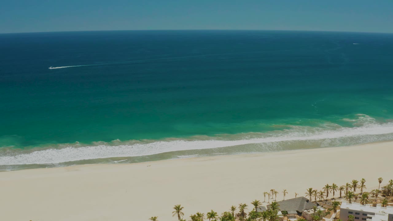 Aerial view of the San Jose del Cabo Estuary coastline as the offshore boat wake sits nearer the center, with turquoise water, white surf, sand, palm trees, roofs, and no visible people.