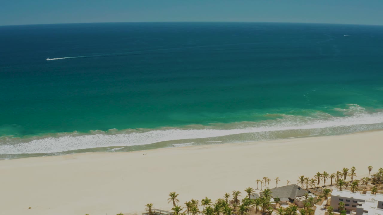 Wide aerial frame over San Jose del Cabo Estuary beach with a boat wake high in the water, foamy surf, broad pale sand, palms and resort buildings along the bottom, and no visible people.