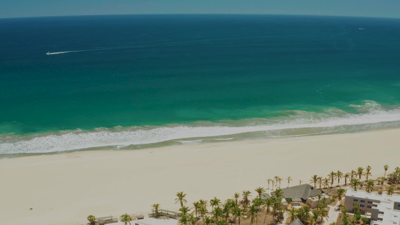 Elevated view of coastline with blue water, breaking waves, empty sand, palms, and buildings.