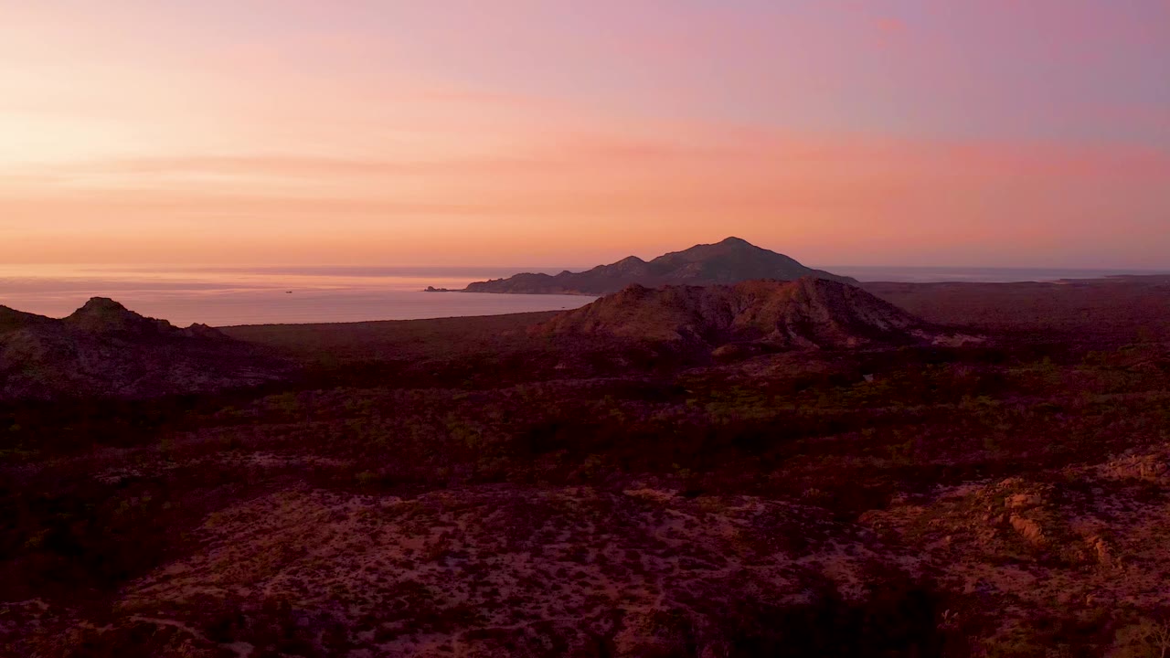 Aerial frame over Cabo Pulmo with dark arid ground in front, a centered mountain ridge, ocean water to the left, and no visible people.