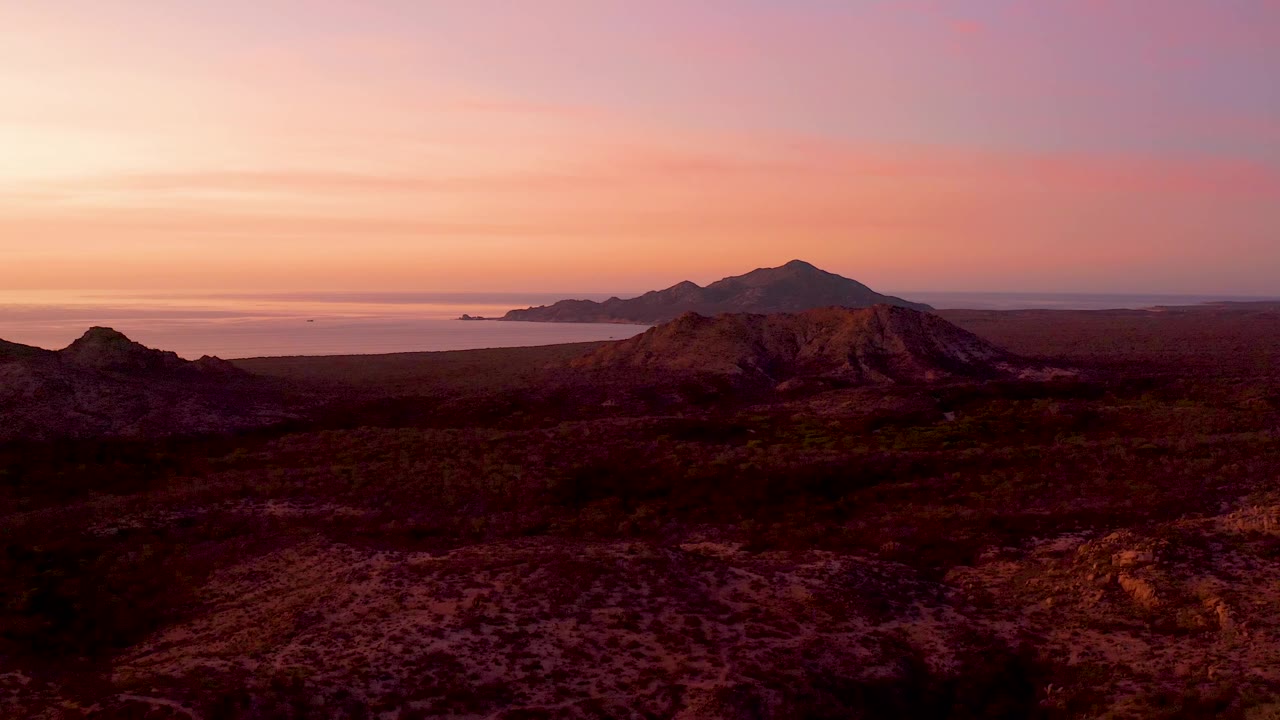 Cabo Pulmo aerial view with a small distant boat on the ocean, layered desert ridges, a central mountain, pink-orange cloud bands, and no visible people.