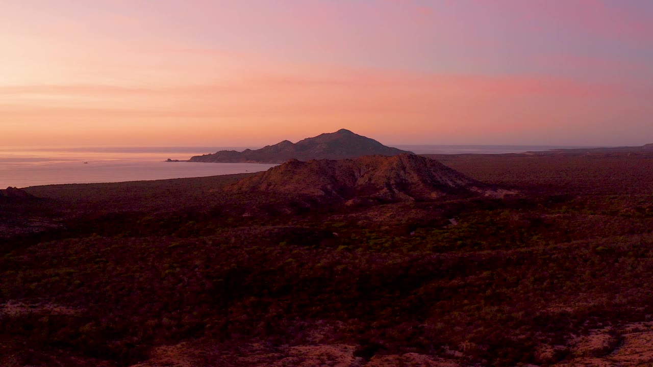 Wide sunset view of a desert hill, ocean, and distant mountains.