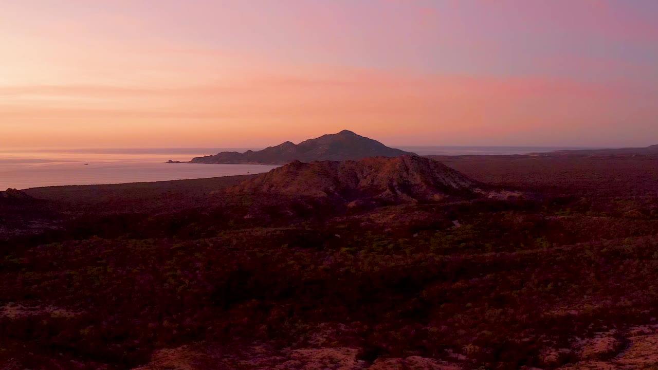 Aerial frame from Cabo Pulmo showing the central rocky hill, dark scrub-covered terrain, ocean to the left, distant coastal mountains, and no visible people.