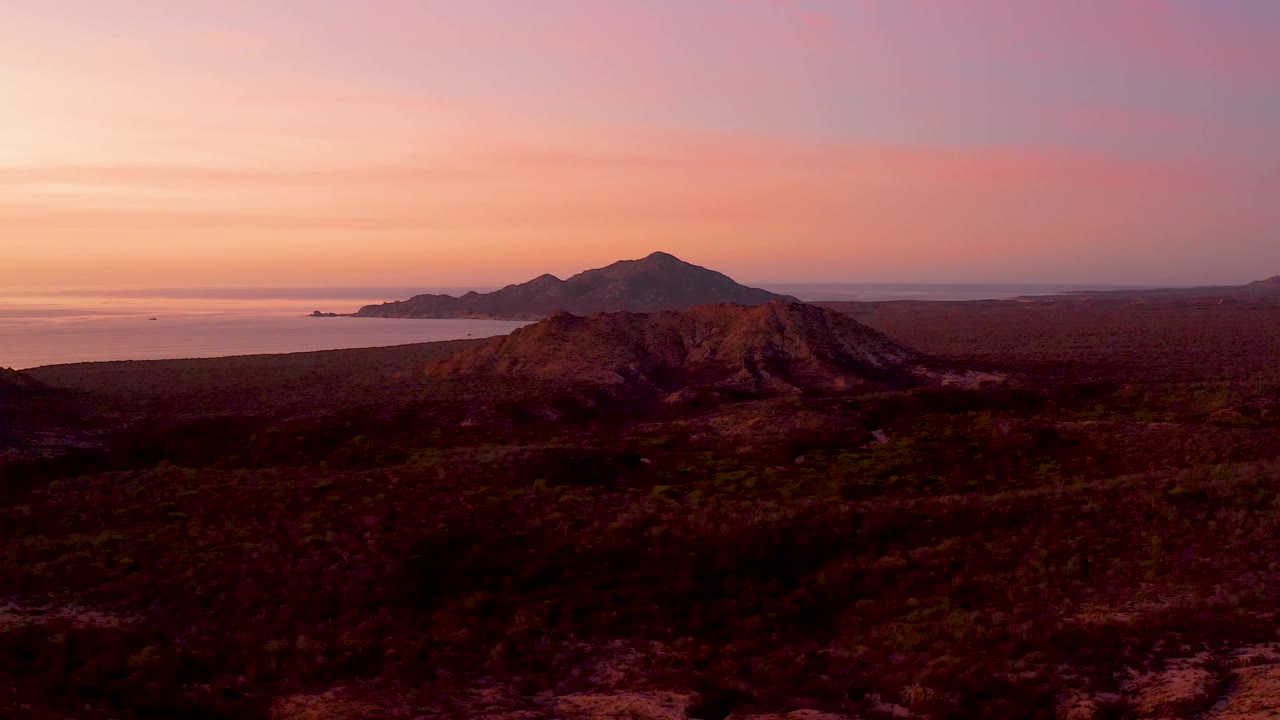 Cabo Pulmo aerial view with a rounded desert hill near center, dark dry ground in front, ocean and low islands or landmasses on the left horizon, and no visible people.