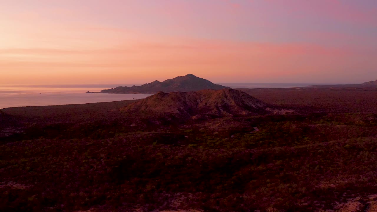 Aerial sunset frame in Cabo Pulmo with the foreground hill slightly lower in the composition, broad dark desert terrain, ocean to the left, distant mountains, and no visible people.