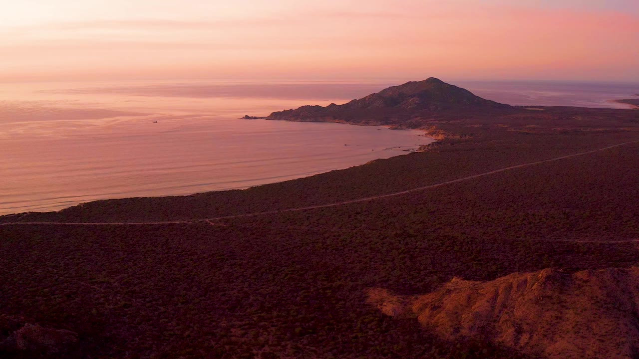 Wide aerial frame in Cabo Pulmo, Baja California Sur showing a coastal road through dark scrubland, ocean at left, a hill on the headland, pink sunset sky, and no visible people.