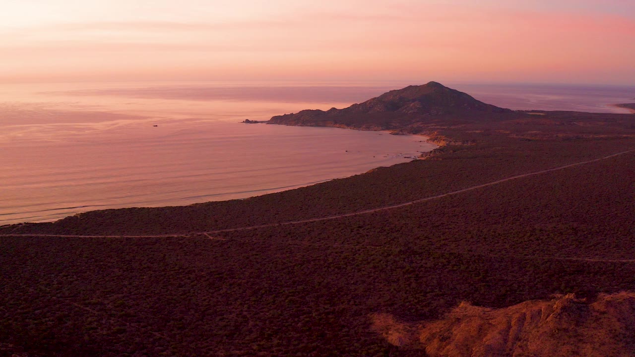 Opening aerial frame over Cabo Pulmo with dark coastal terrain, a road line, calm ocean, distant mountain, pink-orange sky, and no visible people.
